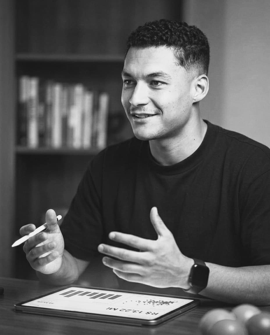 Foto em preto e branco de Matheus explicando uma análise, com um tablet mostrando um dashboard